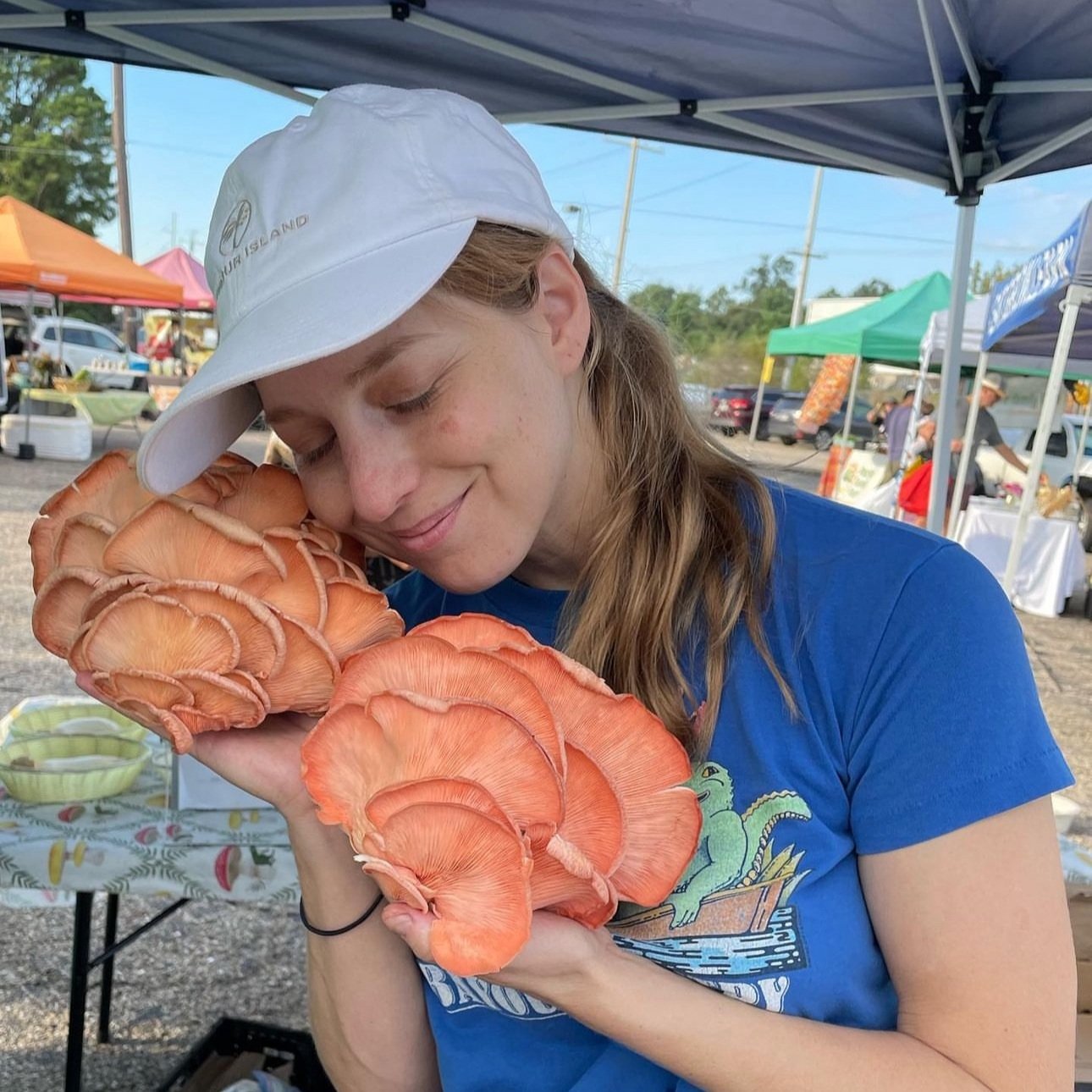 Woman smiling and holding large, vibrant orange mushrooms at a farmers market, showcasing fresh produce and a lively outdoor setting.